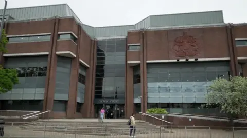 Harrow Crown Court: a large building with brown brick and glass panels. Steps lead up to the entrance of the building with a handful of people outside.
