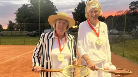 Cirencester Tennis Club Two older women on a clay court dressed up in tennis gear from the olden days - both are wearing long white skirts and one woman has a blue and white striped blazer and straw boater. The other is wearing a head scarf. They are both holding wooden rackets and have large medals on red ribbons round their necks. 