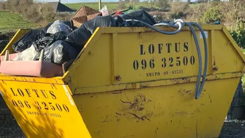 Nicola Byrne A large yellow skip contains several items, including a sofa, a wicker basket, some tubing, and several black bin bags. The skip is situated in a grassy field, with a few houses nearby.
