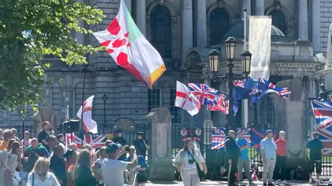 Union jacks are tied to the railings at Belfast City Hall. There are two dozen or so people standing in front of the flags.
