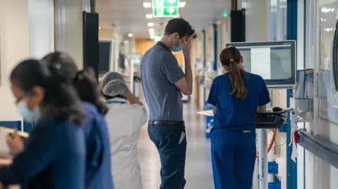 Generic view of two medical staff members from behind looking at a laptop. Some other people in scrubs are seen blurred walking around them.