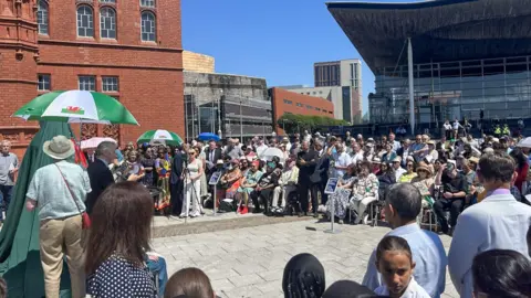 a crowd of people in front of the senedd standing in front of the statue with a green blanket covering it. 