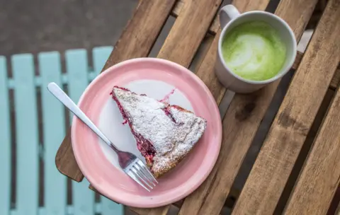 BBC A cherry pie on a pink plate with a fork, and a cup of matcha latte, on a wooden table - photo is taken from above