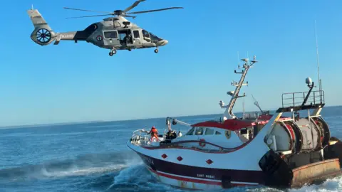 Guernsey Coastguard A helicopter is hovering above a white trawler. A line can just be seen at the front of the boat beside a person wearing a harness and red clothing.