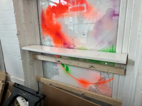 The damaged window from inside the store shows a boarded up window with the remnants of the smashed glass and planks of wood to reinforce the boarding