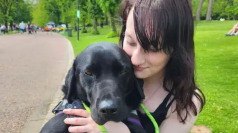 Elizabeth Kane A teenage girl with dark hair cuddles a black Labrador. It is sunny and they are in a park