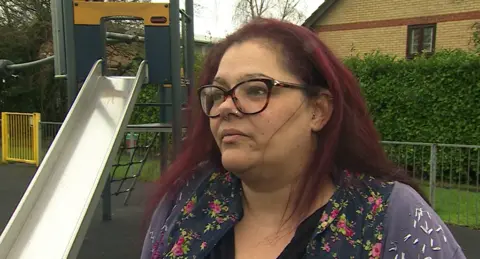 Saffy Charles has purple hair, brown eyes and has glasses on. She is wearing a blue and pink flowery blouse with a purple cardigan. She has a children's playground in the background.