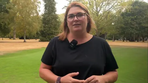 BBC/Lilah Parsons Chelsea Addis wearing glasses and a black top stands on a green area of a parkland golf course, with mature trees and dry grass visible in the background. A clip-on microphone is attached to her top.
