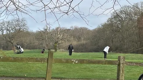BBC The golf course is on the other side of a wooden fence and there are golfers dressed in black and white playing on the pitch. Their trolleys are nearby. The golf course has trees around it and along the hill in the background.