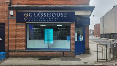 The exterior of Glasshouse Chemist. It is based in a brick building and has a navy blue sign above the main window. The front door, also navy blue, is to the right.
