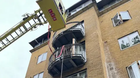 Handout Image showing a yellow turntable ladder with a platform hovering above Mr Rollinson's balcony and attached with a red rope