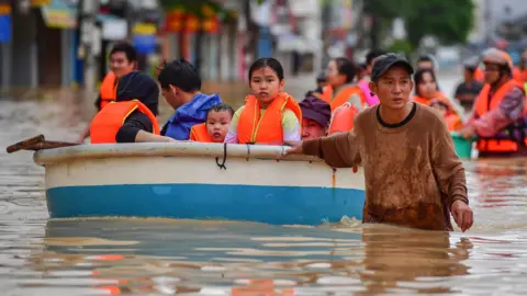 A man wades through floodwater pulling a boat filled with people in life vests.