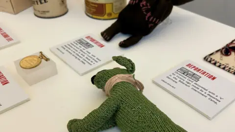 Guernsey Arts There is a white table. On top of it there is signs and knitted animals there.