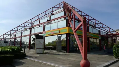 Cameraman/Geograph The Point seen from one corner, with its red pyramid metal bars over the corners of the building. Mirrored glass blocks make up the outer walls and there is a yellow sign over one corner saying Pizza Company. There are three storeys like a square wedding cake, with each smaller than the tier below.