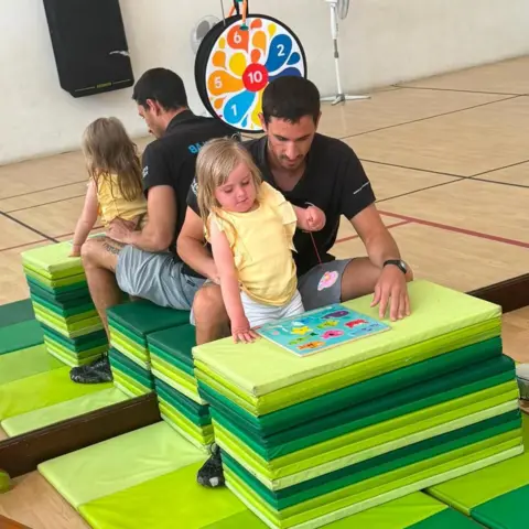 Warren Davies A young blonde girl wearing a yellow t short and white shorts. She is leant up against a stack of green play mats, playing with a coordination jigsaw game in front of her. She is being supported by a man with brown haor, wearing a black polo short and grey shorts. He is sat being her, on a another stack of green mats. Behind them both is a large mirrored wall.