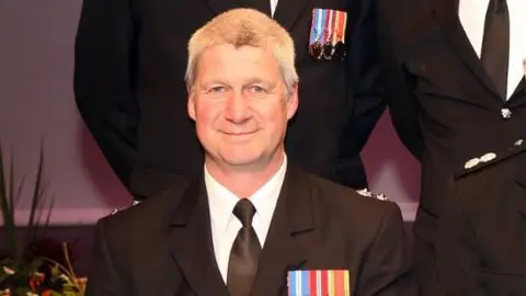 Shropshire Fire and Rescue Service A man with short grey hair and a black suit with medal ribbons on the pocket. Behind him, we can just see the torsos of other men in suits.