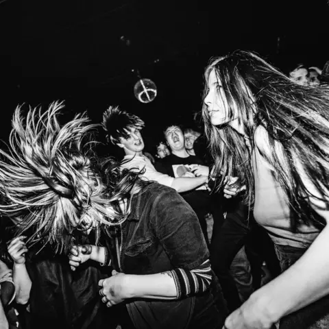 Argiris Liosis A black and white image of Marta del Pinto in a crowd at a gig. Others can be seen dancing beside her
