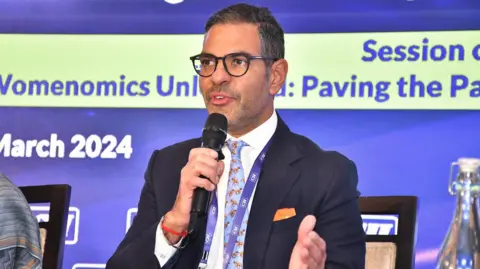 Sunjay Kapur/x Sunjay Kapur wears a black suit with a blue printed tie and a white shirt with a microphone.