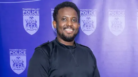 Thames Valley Police PC Ahmed smiles as he stands next to a purple backdrop covered with Thames Valley Police badges. He has short, curly hair and a neat beard and moustache, and wears a black top.