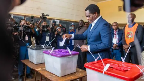 EPA Hakainde Hichilema in a classroom turned polling station places a vote in a ballot box with a purple plastic lid. He is surrounded by lots of people holding cameras.