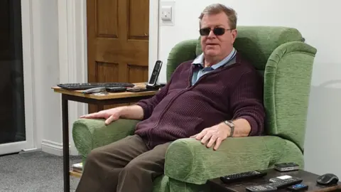 Handout Roger Rees-Evans at home in his armchair surround by technology