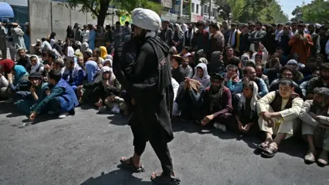 WAKIL KOHSAR/AFP A Taliban fighter walks in front of people sitting along a road outside a bank waiting to withdraw money at Shar-e-Naw neighborhood in Kabul.