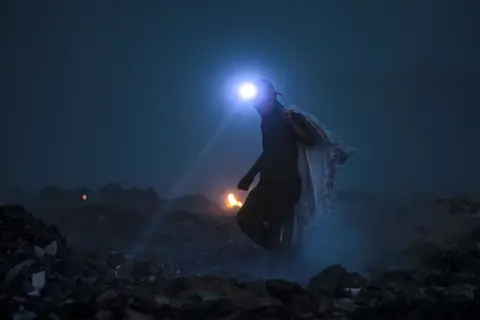 Hussein Faleh / AFP A man equipped with a torch scans for recyclable material at a landfill