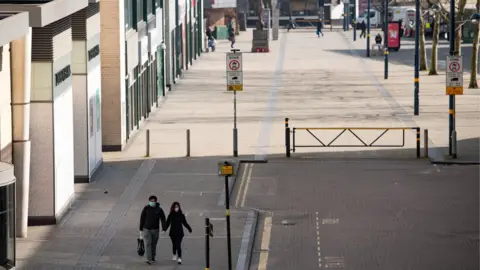 PA Media A couple wearing protective equipment walk by the Bullring in Birmingham, the day after Prime Minister Boris Johnson put the UK in lockdown to help curb the spread of the coronavirus