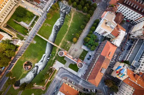 Valentin Flauraud for Saype / Handout via REUTERS A giant land painting of interlocking hands, seen from above
