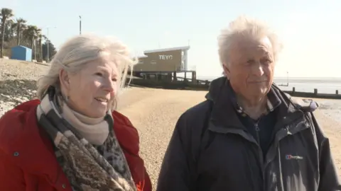 Jamie Niblock/BBC Terri Stanley wearing a red coat and Eric Stanley in a black coat on the beach in Southend