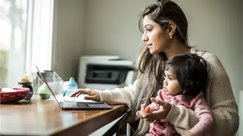 Getty Images Stock image of a mum holding an infant while working on a laptop.