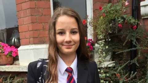 Russell Family 14-year-old Molly Russell is standing in front of a red brick house with pink flowers in a window box in the background. She is wearing her school uniform, a dark blue blazer and red striped tie, smiling at the camera.