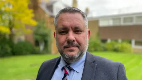 Sam Read/BBC Man with short dark hair and beard wearing a suit and tie and standing outside