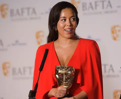 Getty Images Nicôle Lecky wears a red dress. She's standing next to a lectern, answering questions from reporters as she holds the BAFTA award she just won in the Best Miniseries category