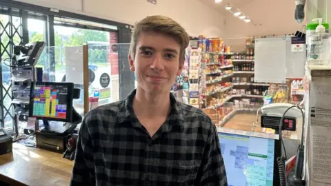 Ollie Conopo/BBC Young man in dark shirt in front of the till of a shop