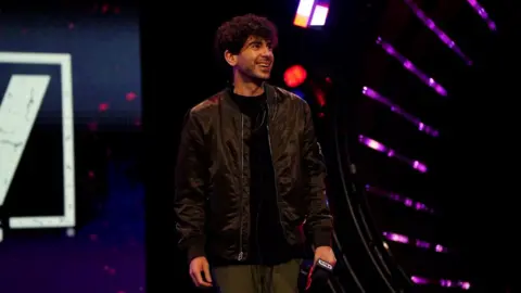 Reuters Tony Khan, co-founder of AEW, pictured on stage. Tony, a man in his 30s, has short curly dark hair and brown eyes. He is wearing green trousers and a black bomber jacket over a black T-shirt. In his left hand he is holding a microphone and is smiling off stage to the right of the camera.