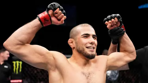 Getty Images Muhammad Mokaev, a man, celebrates his victory over Cody Durden in a flyweight fight. He is smiling and has both arms in the air. He is wearing black fingerless fighting gloves.