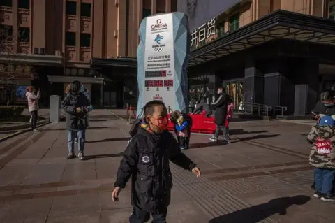 EPA Parents with their children wearing face masks stand next to a countdown display showing days left for the Beijing 2022 Winter Olympics, on a shopping street in Beijing, China, on 17 January 2022