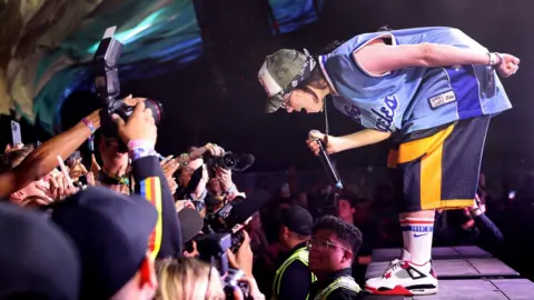 Getty Images Billie Eilish performing at Coachella Festival. Billie is a 22-year-old white woman with shoulder-length dark hair. She is pictured on stage, bending towards the crowd. She wears blue and yellow sports shorts, matched with a blue sports jersey, white sports socks and white sneakers with red trim. She wears a green and white cap over a blue bandana and holds the microphone in her right hand while her left is behind her back. A line of security officers separates her from the crowd, which is otherwise very close. People in the crowd hold phones and cameras up to capture her performance.