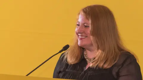 Neil Harrison  Naomi Long smiles as she speaks into a microphone against a yellow background