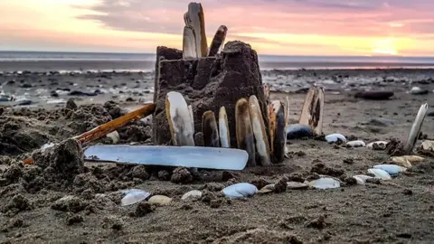 Ashley Williams Sandcastle in foreground including shells with low tide line in distance and orange and yellow sky