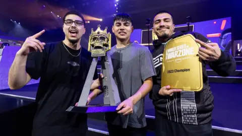 WSOW Three young men celebrate winning the World Series of Warzone global final. They show off the competition trophy - a golden skull, with a crown, on a tripod. One of the men also holds a gold plaque with "World Series of Warzone: 2023 Champion" written on it.