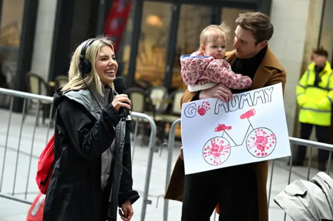 Jeff Spicer/Comic Relief A woman holding a radio microphone and wearing large over-ear headphones smiles widely as she looks lovingly at a tall man with short brown hair who's holding a young child in his arms. The man is also holding a large white sign with "Go Mummy!" and a picture of a bicycle on it.