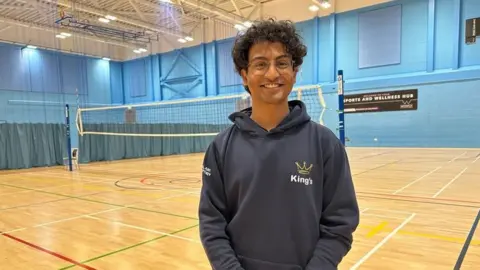 A young man wearing glasses stands in front of a volleyball net in an indoor sports court. The walls are painted light blue and cricket nets can be seen at the far end of the court. A black sign reading "sports and wellness hub" is stuck on the wall.