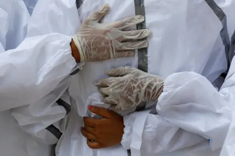 Adnan Abidi / Reuters Relatives wearing personal protective equipment (PPE) mourn a man and place hands on each other's backs