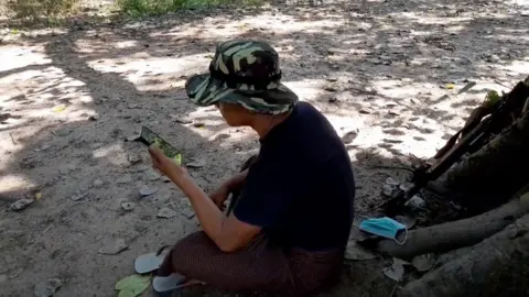 BBC Bo Kyar Yine seated in a clearing under a banyan tree on the phone to his son