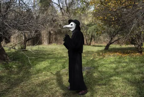 Ihsaan Haffejee/ Getty Images Man wearing medieval clothing - a long black outfit with a white bird-like mask. He is standing in what looks like a garden - 3 June.
