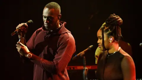 Getty Images Stormzy and Debbie perform together onstage. Stormzy is wearing a red hoodie, in a ribbed texture. He holds the microphone with both hands. Debbie wears her hair up, she's wearing a sparkling necklace and smiling, looking out to the audience.