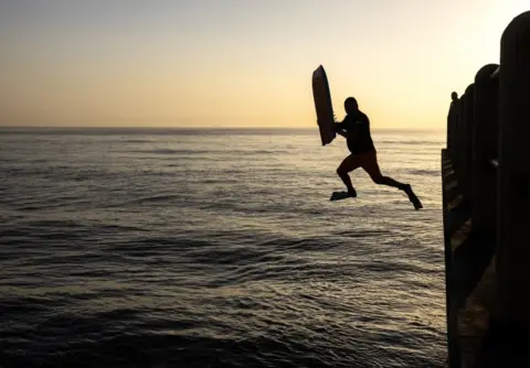 KIM LUDBROOK/EPA A surfer jumps off the pier in the early morning as the sun rises above North Beach Pier in Durban.