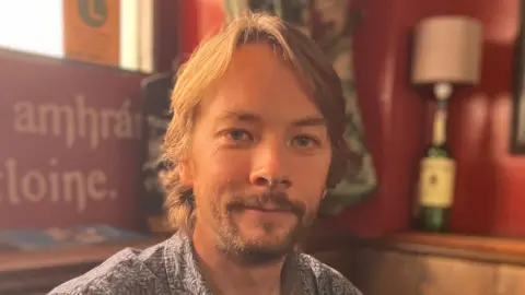 Conor Lamb who has medium length brown hair and a brown goatee/beard. He is wearing a blue and white patterned shirt sitting in a traditional old pub with red walls and old signs in Irish. 